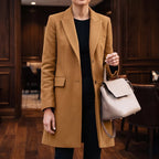 Woman wearing a fitted tan wool knee-length coat over a black top and pants, holding a light gray handbag.