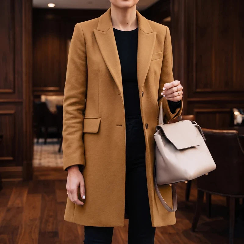 Woman wearing a fitted tan wool knee-length coat over a black top and pants, holding a light gray handbag.
