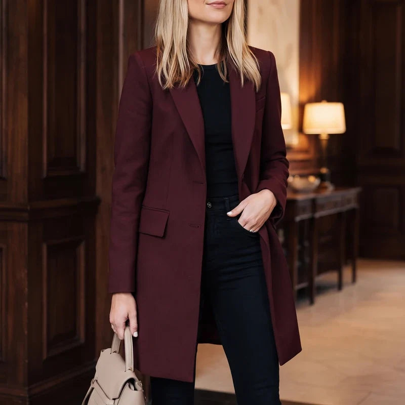 Woman wearing a long burgundy blazer over a black top and pants, holding a beige handbag in a wood-paneled room.