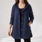 Woman wearing a navy blue quilted hooded jacket with a braided texture, wooden buttons, and pockets, against a light gray background.