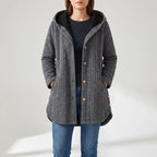 Woman wearing a gray quilted hooded jacket with black lining, wooden buttons, and blue jeans, standing with hands in pockets.