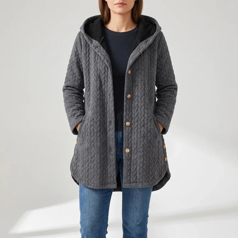 Woman wearing a gray quilted hooded jacket with black lining, wooden buttons, and blue jeans, standing with hands in pockets.