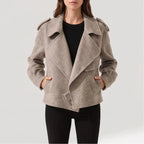 Woman wearing a light taupe short jacket with wide lapels, shoulder tabs, and hands in front pockets, over black pants.
