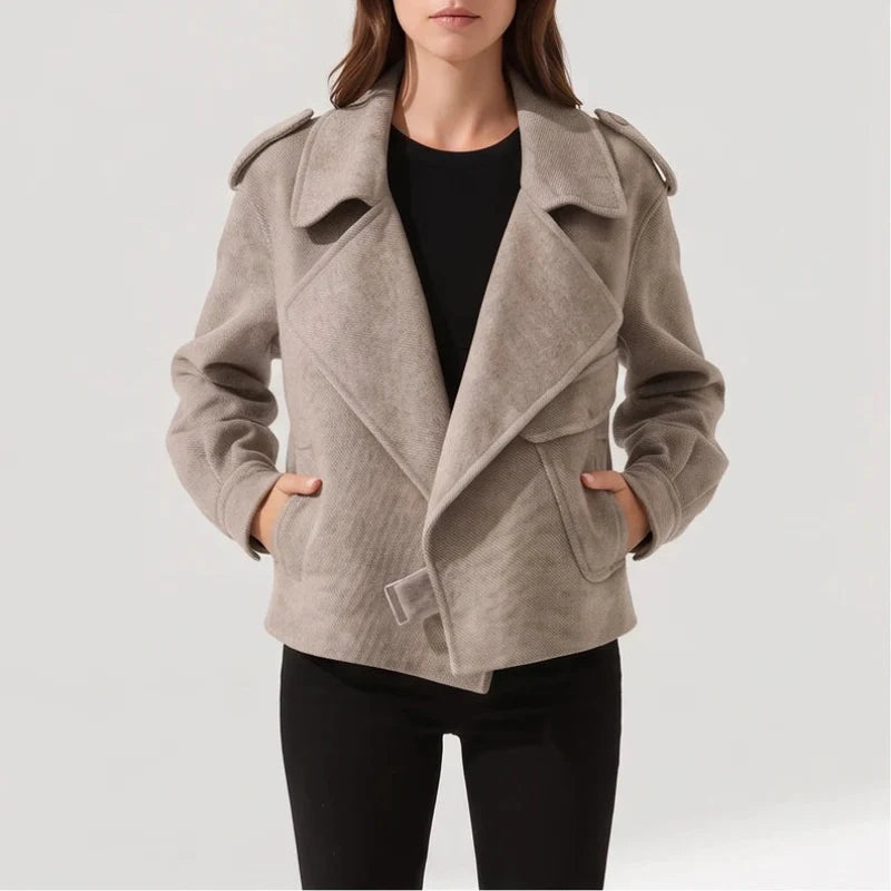 Woman wearing a light taupe short jacket with wide lapels, shoulder tabs, and hands in front pockets, over black pants.