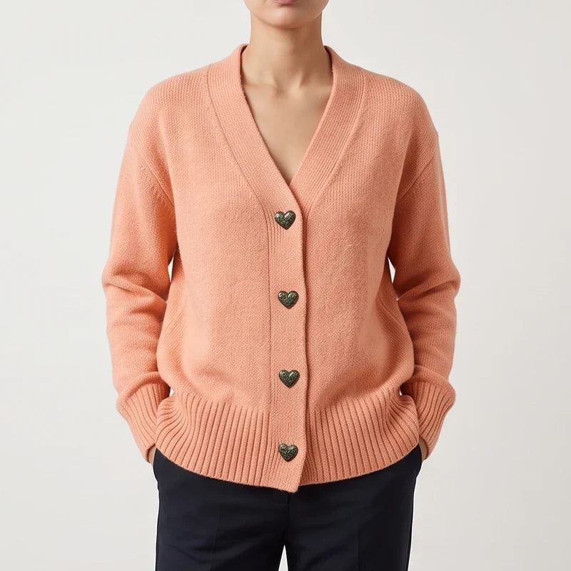 Woman wearing a relaxed-fit, peach knit cardigan with V-neckline and decorative dark heart-shaped buttons, hands in pockets.