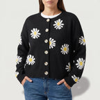 Woman wearing a black knit cardigan with white and yellow daisy patterns, light-colored buttons, and blue jeans.