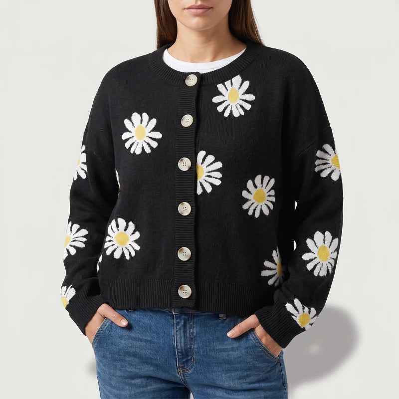 Woman wearing a black knit cardigan with white and yellow daisy patterns, light-colored buttons, and blue jeans.