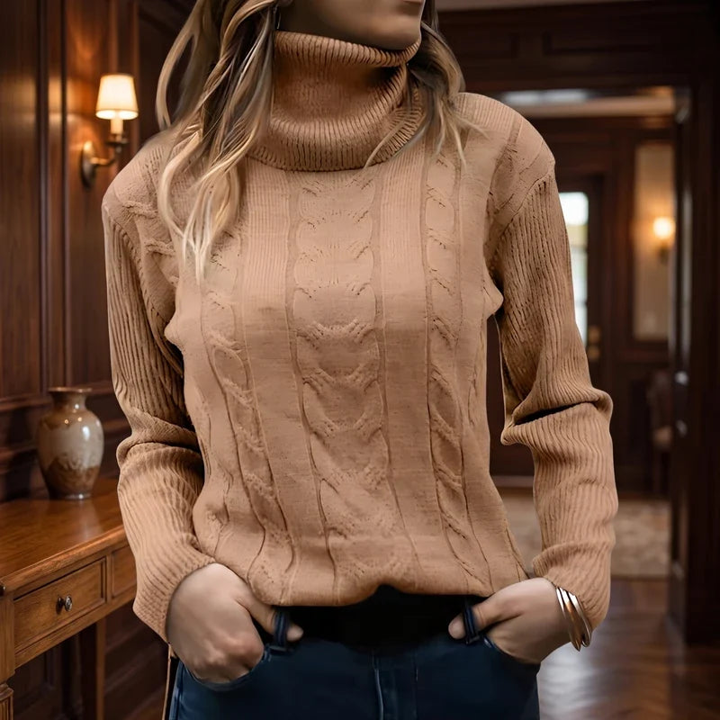 Woman wearing a fitted light brown cable knit turtleneck sweater with ribbed cuffs, hands in dark blue jeans pockets, against a wooden hallway background.