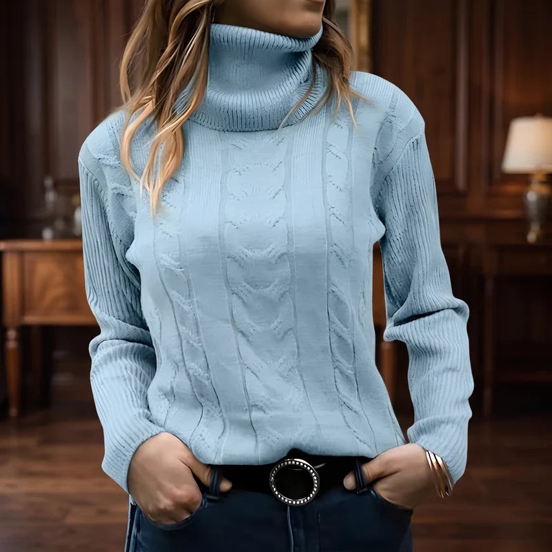 Woman wearing a light blue cable knit turtleneck sweater with ribbed cuffs and collar, hands in jeans pockets.
