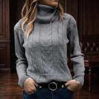 Woman wearing a fitted grey cable-knit turtleneck sweater with ribbed sleeves and a black belt, hands in pockets.