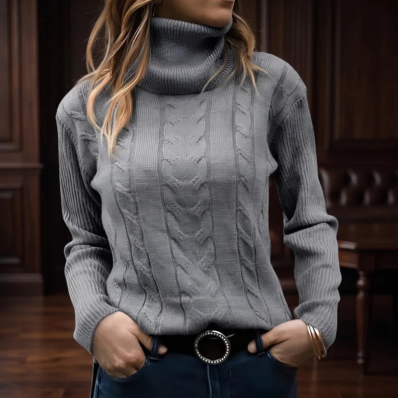Woman wearing a fitted grey cable-knit turtleneck sweater with ribbed sleeves and a black belt, hands in pockets.