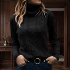 Woman wearing a black cable knit turtleneck sweater with ribbed cuffs and collar, hands in jeans pockets, in a warm wooden room.