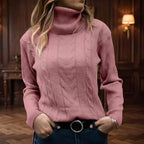 Woman wearing a pink cable knit turtleneck sweater with ribbed cuffs and a black belt, hands in pockets.