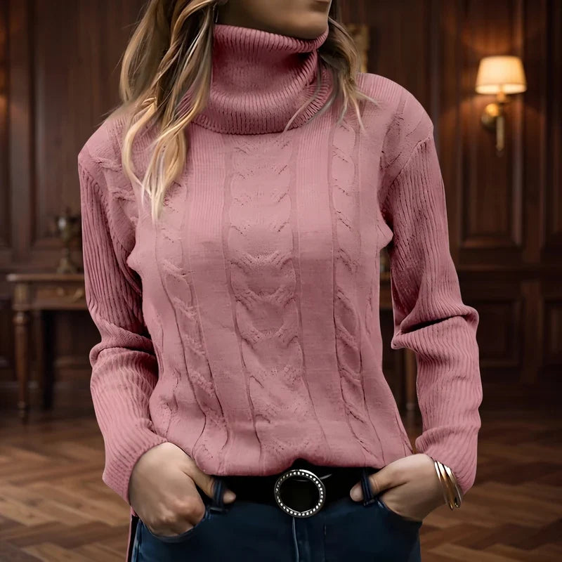 Woman wearing a pink cable knit turtleneck sweater with ribbed cuffs and a black belt, hands in pockets.