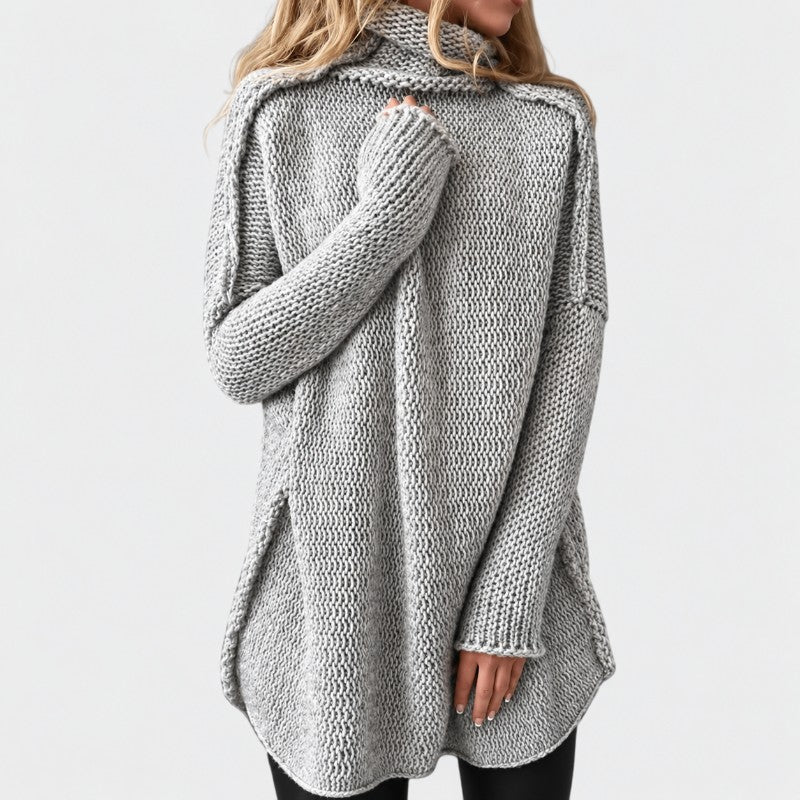 Woman wearing a light grey, chunky knit tunic sweater with a high rolled neck, long sleeves, and a curved hem with side slits.
