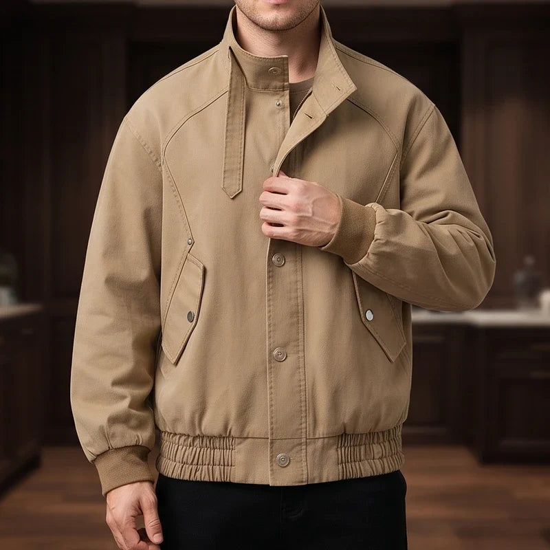 Man wearing a tan cotton jacket with a stand collar, button placket, ribbed cuffs and hem, hand in pocket.