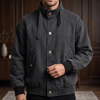 Man wearing a dark charcoal gray jacket with a high buttoned collar, flapped pockets, and ribbed cuffs.