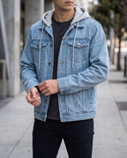 Man wearing a light wash denim jacket with a grey hooded sweatshirt layered underneath, adjusting his cuff.