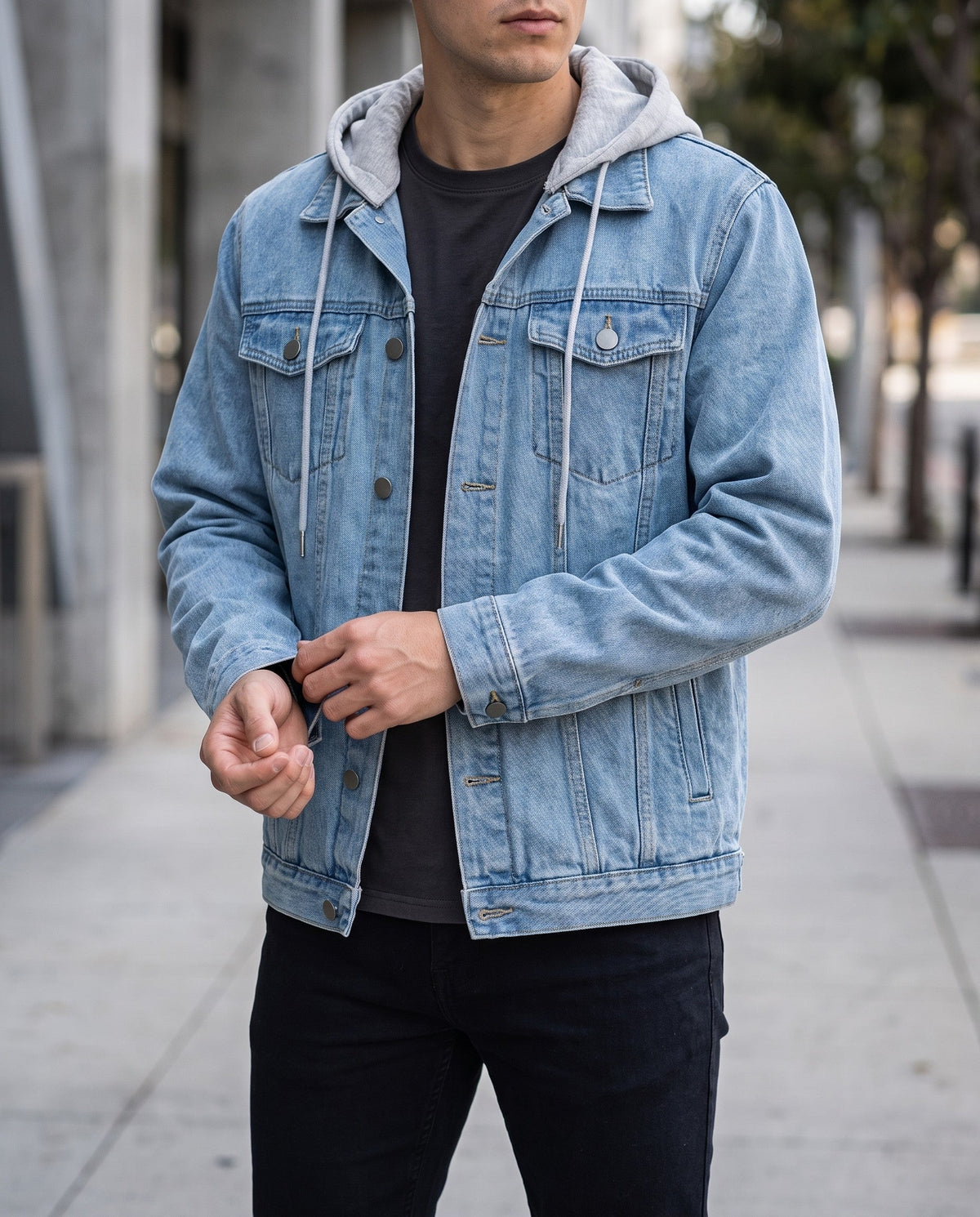 Man wearing a light wash denim jacket with a grey hooded sweatshirt layered underneath, adjusting his cuff.