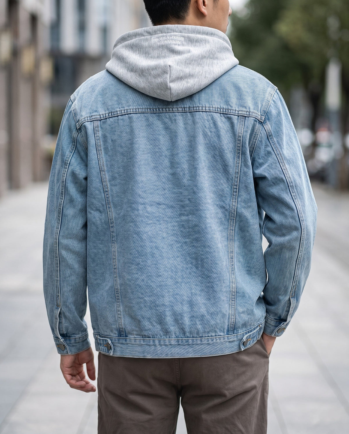 Man wearing a light blue denim jacket over a gray hooded sweatshirt, standing with his back to the viewer outdoors.