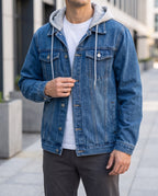 Man wearing a blue denim jacket with a grey hooded sweatshirt underneath, buttoning the front over dark pants.