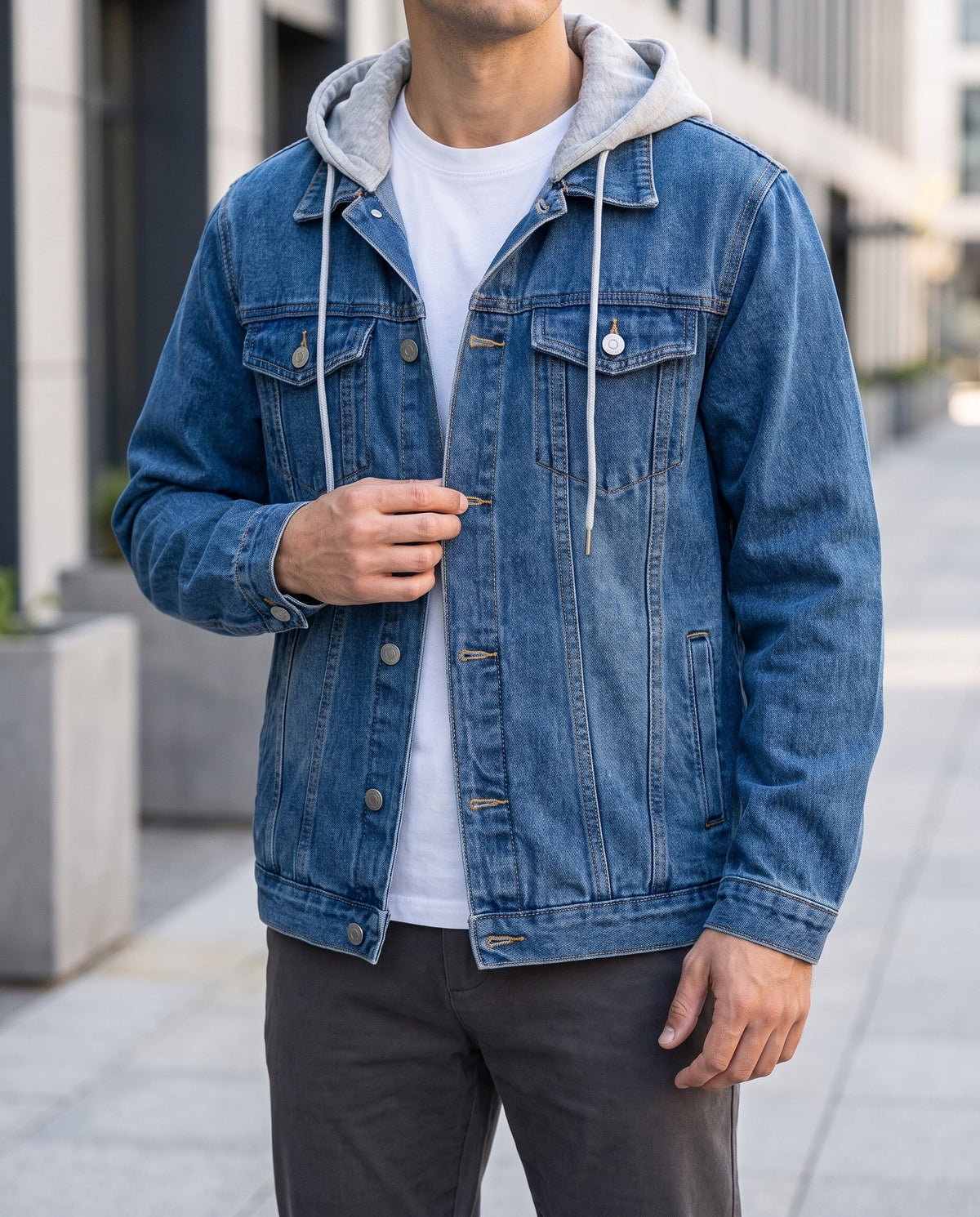Man wearing a blue denim jacket with a grey hooded sweatshirt underneath, buttoning the front over dark pants.