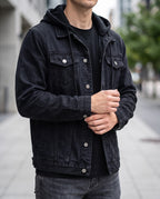Man wearing a black distressed denim jacket over a black hooded shirt, hands clasped, outdoors on a street.
