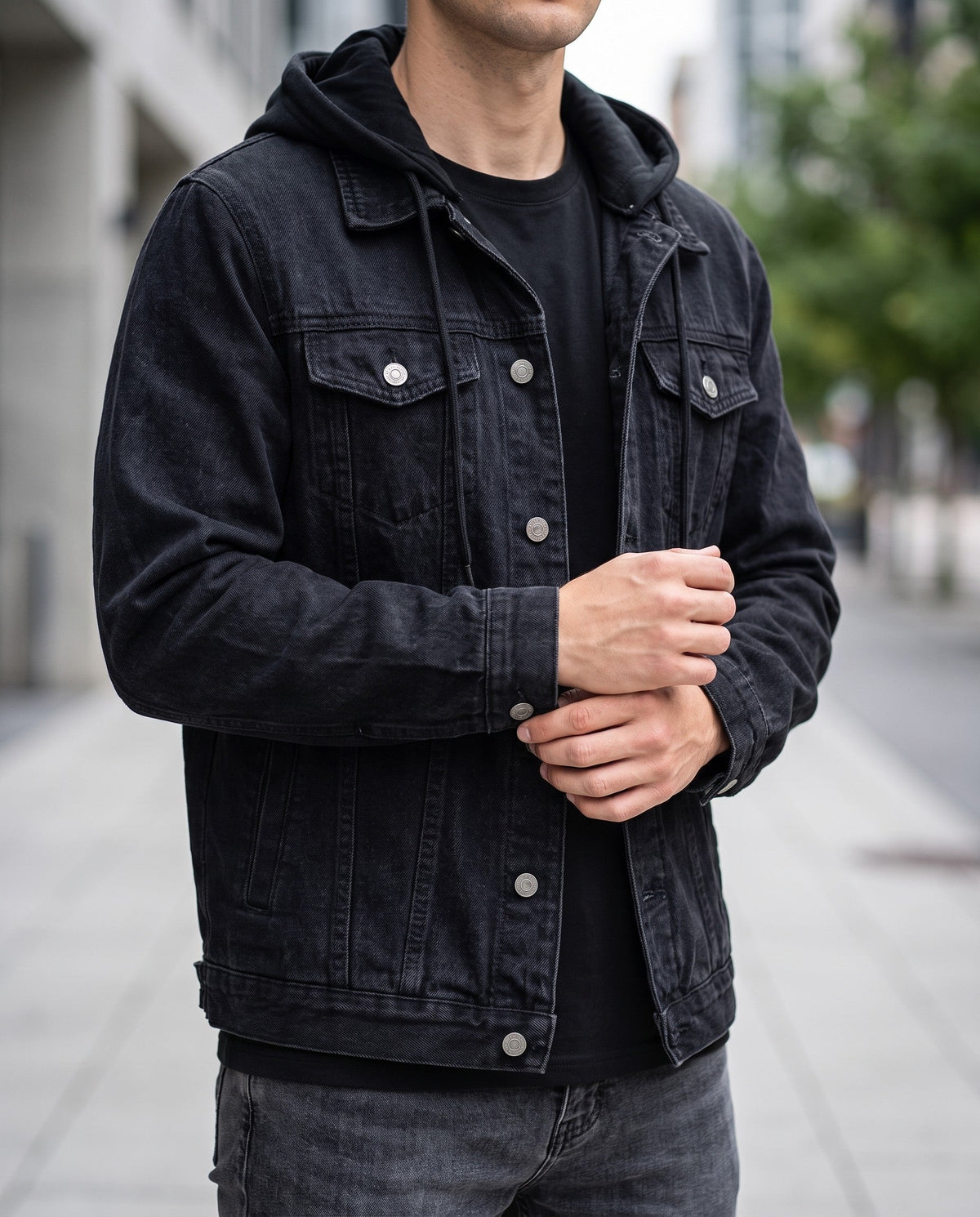 Man wearing a black distressed denim jacket over a black hooded shirt, hands clasped, outdoors on a street.