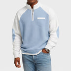 Man wearing a light blue and white color-blocked waffle knit half-zip sweatshirt with a chest pocket and light wash jeans.