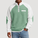 Man wearing a sage green and white color-block waffle knit half-zip sweatshirt with a chest pocket and elbow patches.