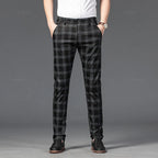 Man wearing black and gray plaid straight-leg trousers with a white shirt, hands in pockets, standing on a dark background.