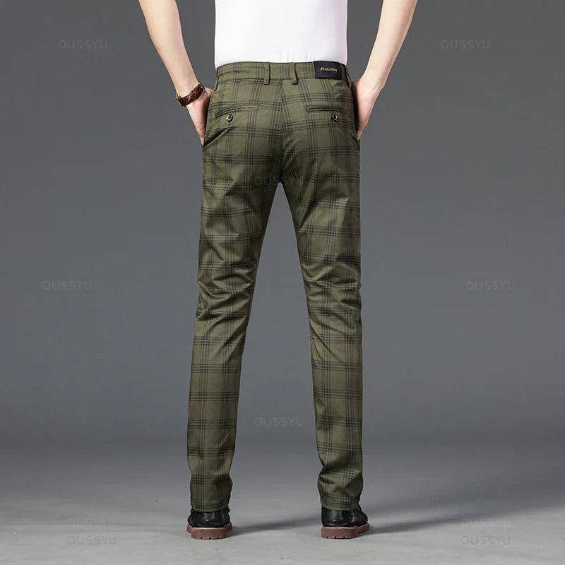 Man wearing olive green plaid straight-leg trousers with back buttoned pockets, hands in front pockets, standing against a dark grey background.