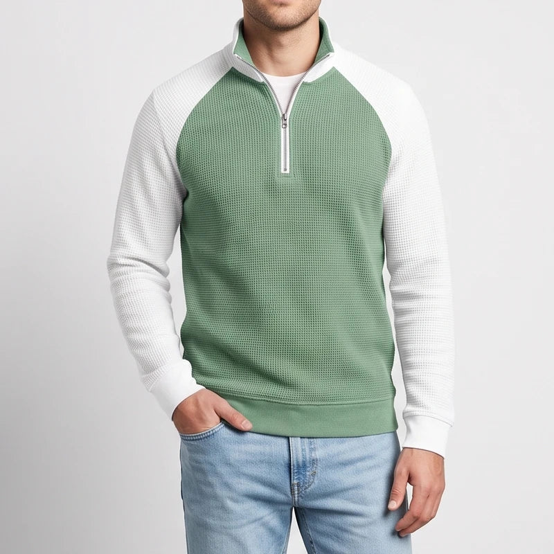 Man wearing a two-tone green and white waffle-knit quarter-zip sweatshirt with light blue jeans, hand in pocket.