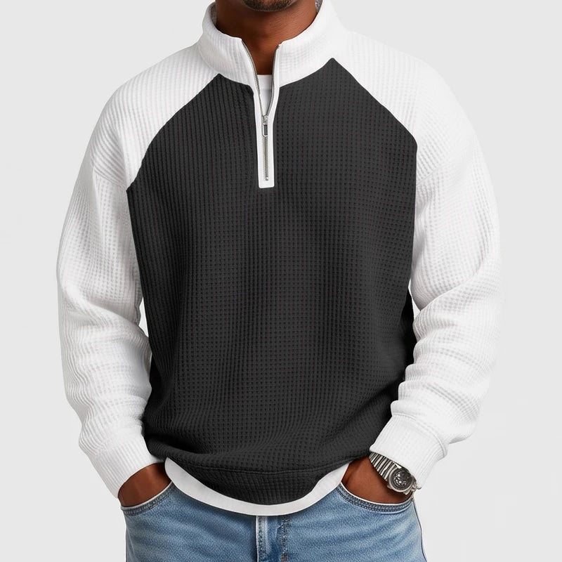 Man wearing a black and white color-block waffle knit quarter-zip sweatshirt with raglan sleeves and a silver zipper, hands in jeans pockets.