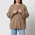Woman wearing a beige chunky knit cardigan with gold buttons, unbuttoning it over light blue jeans.