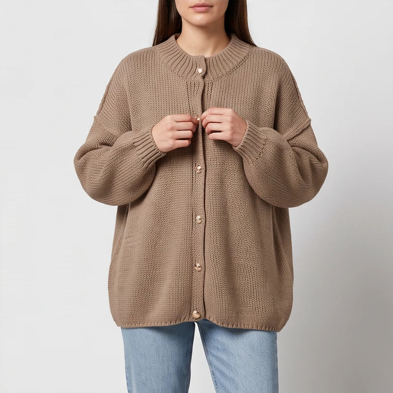 Woman wearing a beige chunky knit cardigan with gold buttons, unbuttoning it over light blue jeans.