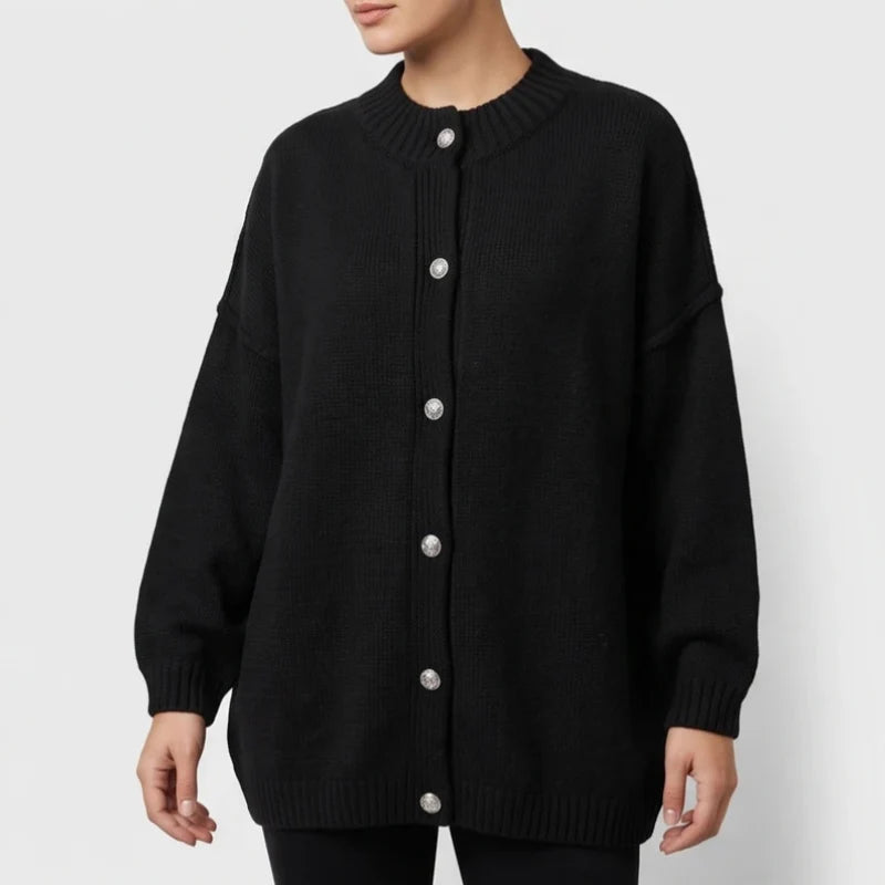 Woman wearing a black oversized knit cardigan with a ribbed collar and cuffs, featuring five large silver-toned buttons down the front.