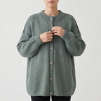 Woman adjusting the ornate silver buttons on a long, loose-fit, muted green knit cardigan with ribbed collar and cuffs.