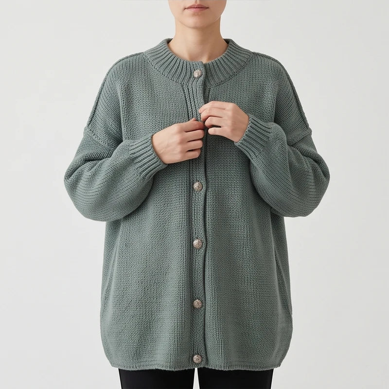 Woman adjusting the ornate silver buttons on a long, loose-fit, muted green knit cardigan with ribbed collar and cuffs.