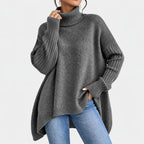 Woman wearing a dark grey oversized knit turtleneck sweater with wide ribbed cuffs and a high-low hem, paired with blue jeans.