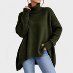 Woman wearing an oversized dark green knit turtleneck sweater with ribbed cuffs and a high-low hem over blue jeans.