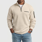 Man wearing a light beige waffle-knit quarter-zip pullover with a black zipper, chest pocket, and sleeve pocket, hands in blue jeans pockets.