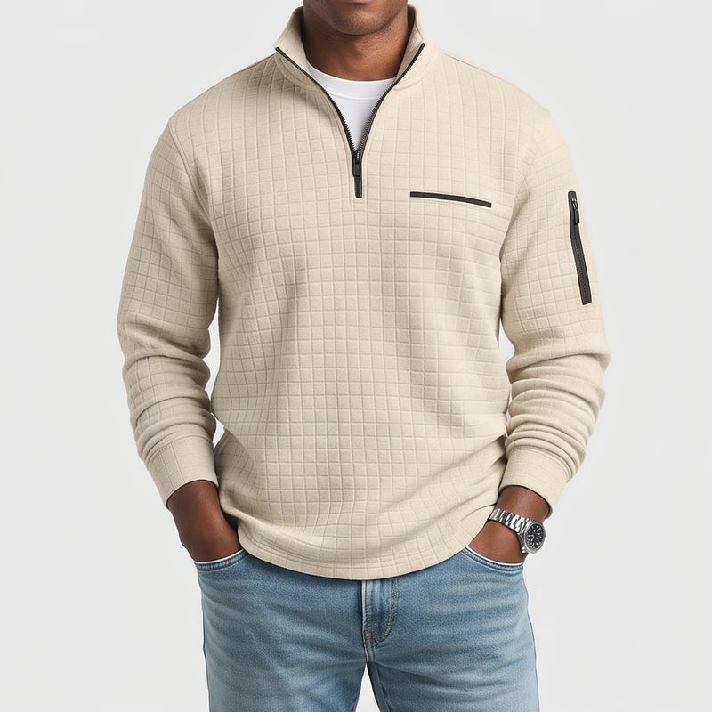 Man wearing a light beige waffle-knit quarter-zip pullover with a black zipper, chest pocket, and sleeve pocket, hands in blue jeans pockets.