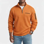 Man wearing an orange quarter-zip waffle knit jumper with black chest and arm zipper pockets, paired with blue jeans.