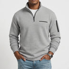 Man wearing a light gray quarter-zip waffle knit jumper with black zippers, chest pocket, and arm pocket, hands in jeans pockets.