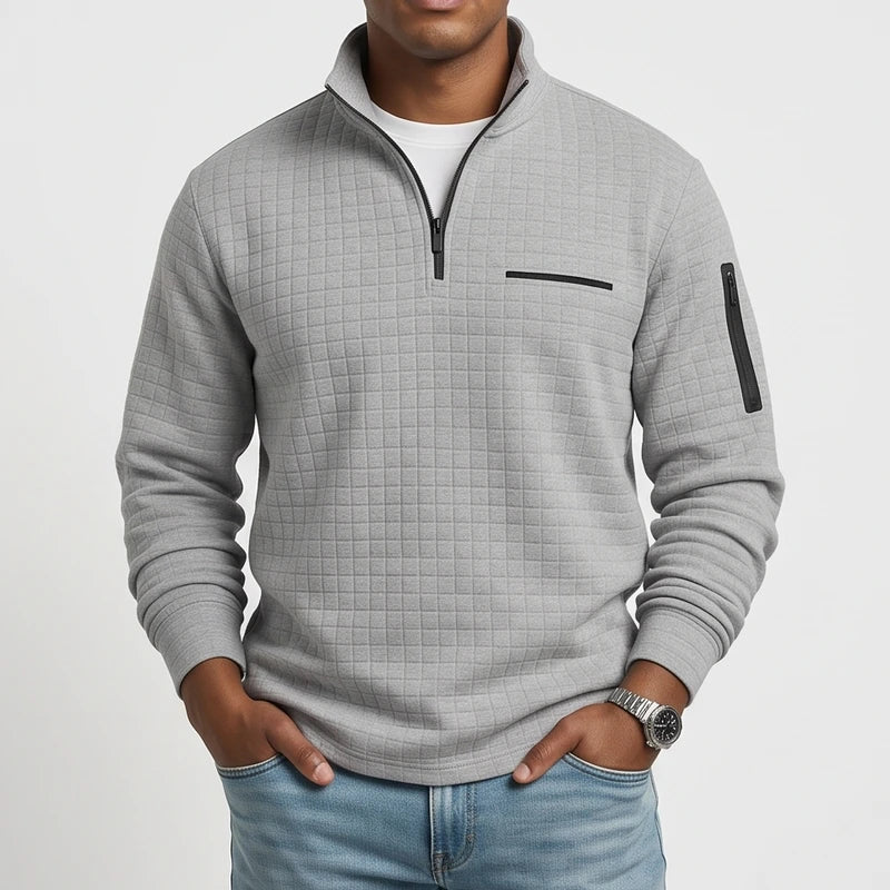 Man wearing a light gray quarter-zip waffle knit jumper with black zippers, chest pocket, and arm pocket, hands in jeans pockets.