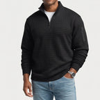 Man wearing a black quarter-zip jumper with a quilted square texture, an arm pocket, and a chest pocket, paired with light blue jeans.
