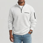 Man wearing a light gray quarter-zip waffle knit jumper with black zippers, a chest pocket, and blue jeans.