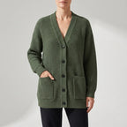 Woman wearing a long olive green chunky knit cardigan with a V-neck, black buttons, and two front pockets.