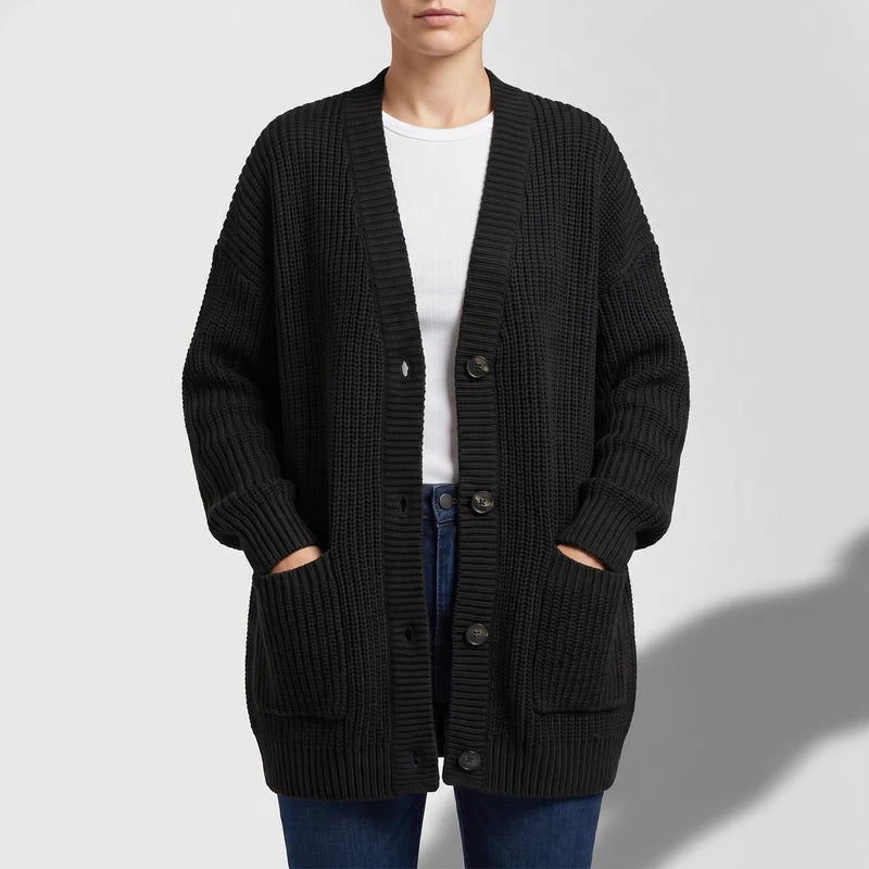 Woman wearing an open black chunky knit cardigan with large brown buttons and front pockets over a white top.